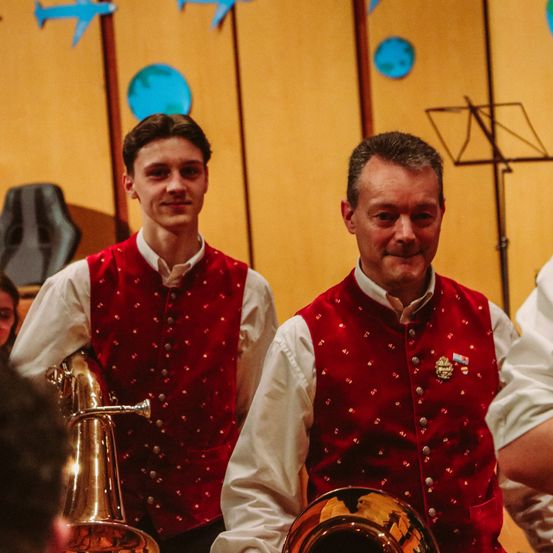 Zwei Männer in roten Westen, einer hält ein Tenorhorn und der andere eine Tuba, stehen in einem Raum mit einer gelben Wand, einem schwarzen Notenständer und blauen Kreisen an der Wand.