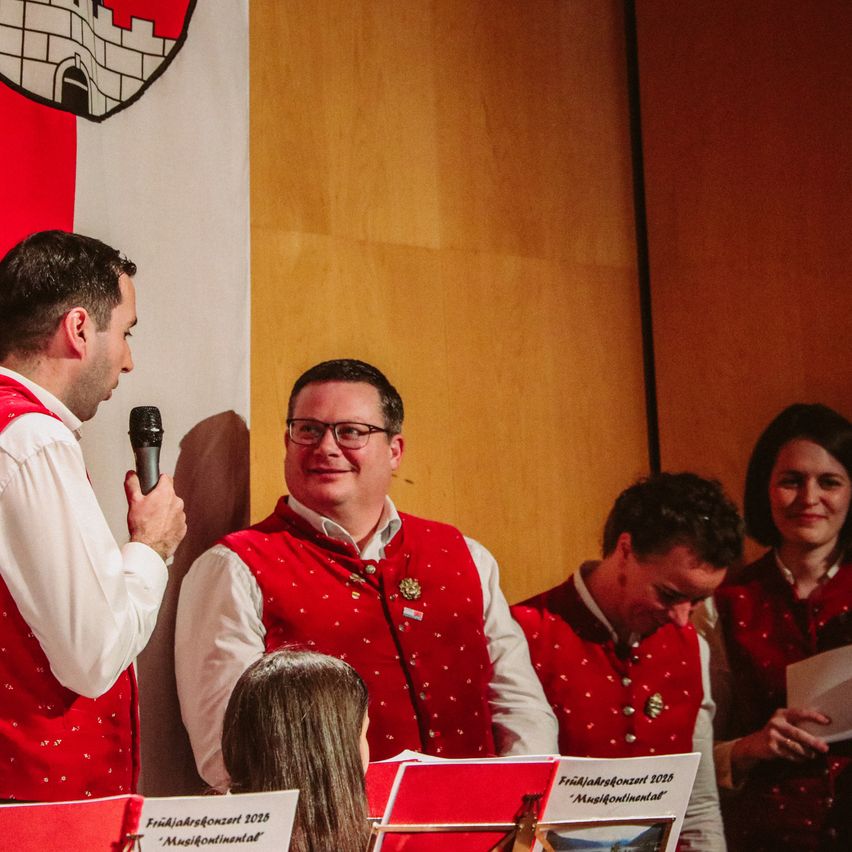 Ein Mann, der ein Mikrofon hält, steht hinter einer Gruppe von Menschen, die rote Westen tragen. Sie halten alle ein Buch und ein Stück Papier in der Hand. Sie treten wahrscheinlich bei einem Konzert auf. Hinter ihnen ist eine Holzwand mit einer Flagge. Die Flagge hat ein Schlossdesign. Der Mann vorne schaut etwas an. Die Leute hinter ihm schauen ebenfalls etwas an. Das Licht ist auf sie gerichtet.