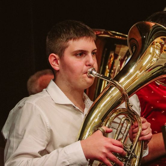 Ein junger Mann in einem weißen Hemd spielt eine Tuba und schaut nach unten, während ein anderer Mann in einem roten Hemd hinter ihm steht.