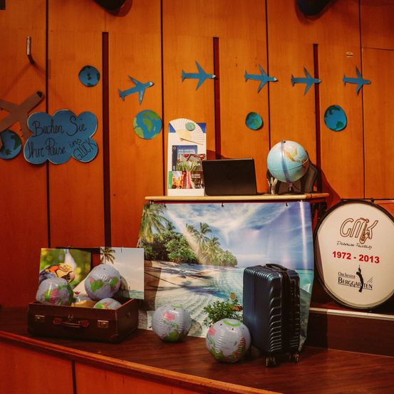 Ein hölzerner Raum mit einem Schreibtisch hat Reiseartikel darauf. Ein Laptop, eine Weltkugel und eine Trommel stehen auf dem Schreibtisch. Es gibt auch einige Koffer und eine Kiste mit Kugeln.