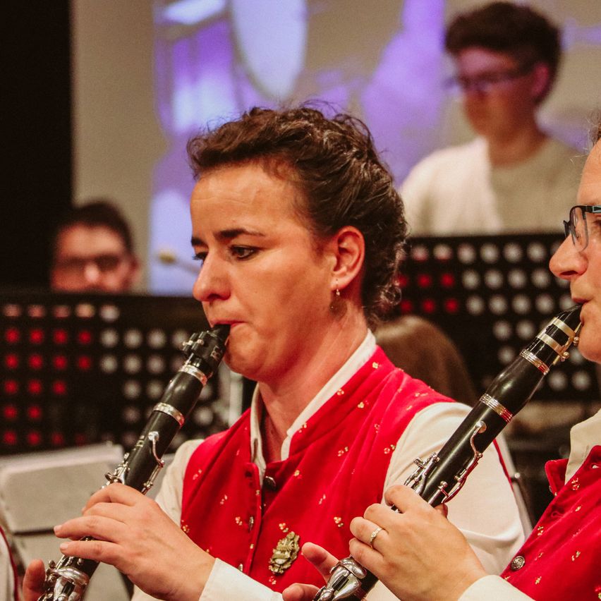 Zwei Frauen in roten Westen spielen in einem Konzert die Klarinette. Dahinter schaut ein Junge in einem weißen Hemd auf eine Projektionsleinwand.