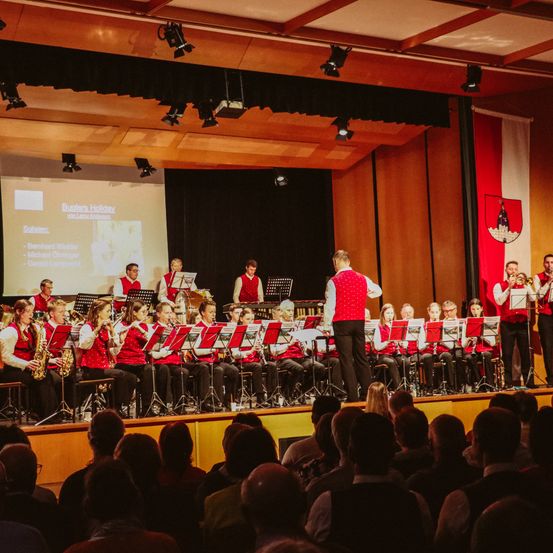 Ein großes Blechbläserensemble spielt auf der Bühne, während eine Projektionsleinwand ihren Namen und eine Flagge im Hintergrund anzeigt. Das Publikum sitzt vor der Bühne und beobachtet die Aufführung.
