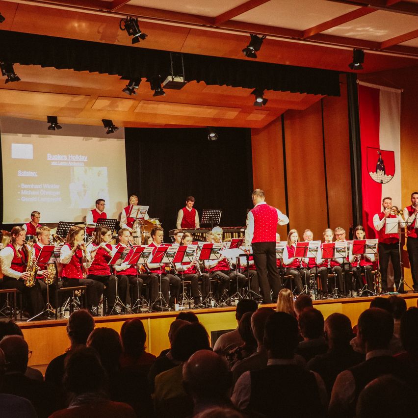 Ein großes Blechbläserensemble spielt auf der Bühne, während eine Projektionsleinwand ihren Namen und eine Flagge im Hintergrund anzeigt. Das Publikum sitzt vor der Bühne und beobachtet die Aufführung.