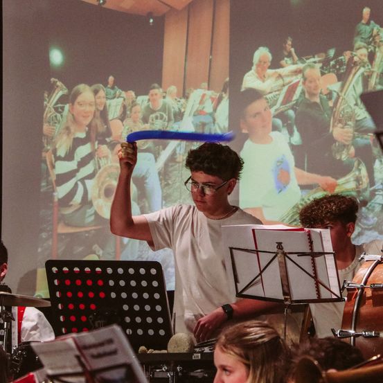 Ein Junge mit Brille spielt ein blaues Spielzeuginstrument, während eine Band von Menschen hinter ihm spielt.