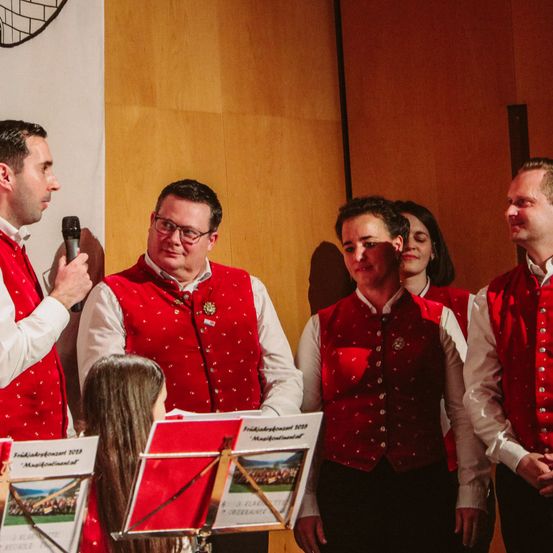 Eine Gruppe von Männern in roten Westen und weißen Hemden steht vor einer Frau, die Geige spielt. Die Männer schauen die Frau an, während sie spielt.