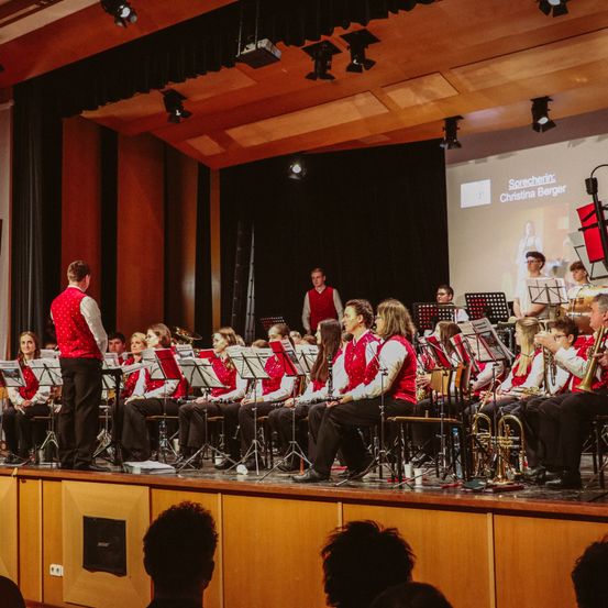 Eine Gruppe von Musikern auf der Bühne, gekleidet in rote Westen, spielt für ein Publikum in einem Theater. Ein Dirigent steht vorne, und eine Projektionsleinwand befindet sich dahinter.