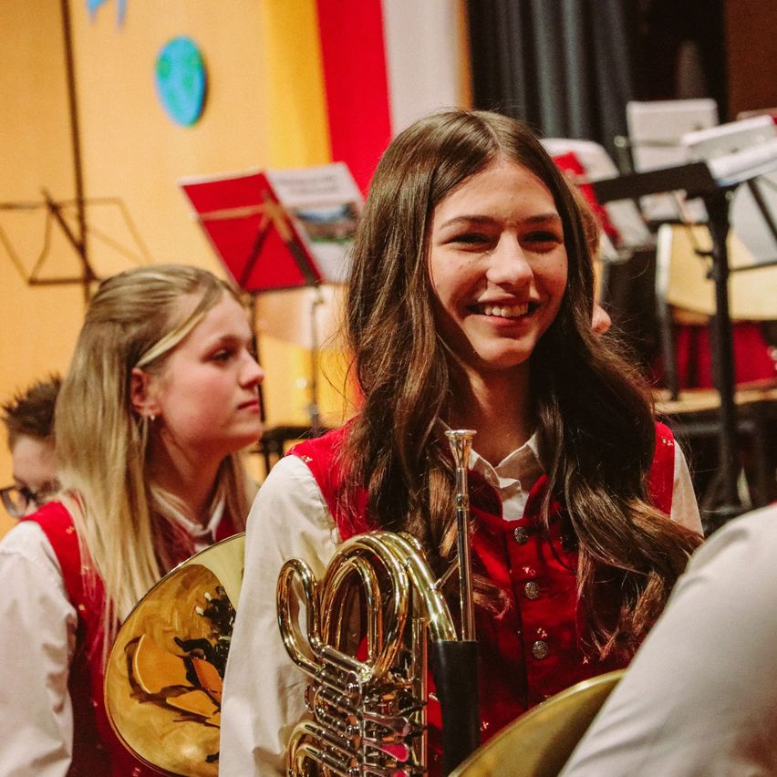 Zwei Mädchen in roten Westen lächeln und halten ein Blechblasinstrument, mit Notenständern und einer Flagge im Hintergrund.
