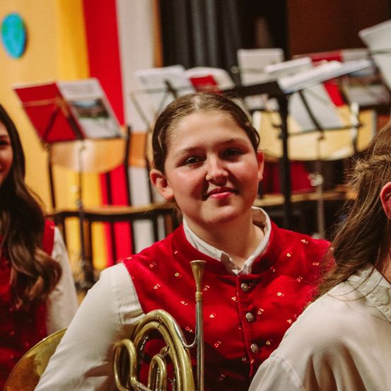 Ein Mädchen in einer roten Weste mit goldenen Knöpfen spielt ein Messing-Waldhorn. Hinter ihr stehen zwei weitere Mädchen, die ähnlich gekleidet sind. Sie befinden sich in einem Raum mit mehreren Musikinstrumenten auf Ständern.