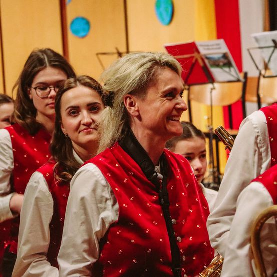 Eine Frau mit grauen Haaren und einer roten Weste mit goldenen Punkten lächelt vor einer Gruppe junger Mädchen, die dieselbe Weste tragen.
