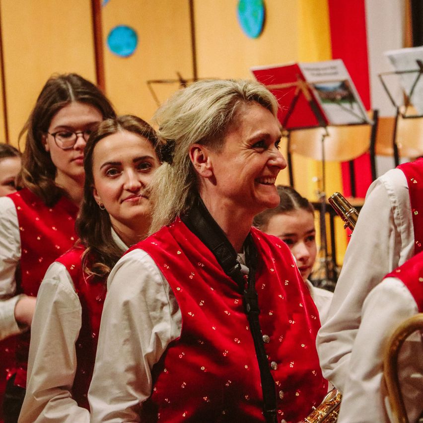 Eine Frau mit grauen Haaren und einer roten Weste mit goldenen Punkten lächelt vor einer Gruppe junger Mädchen, die dieselbe Weste tragen.