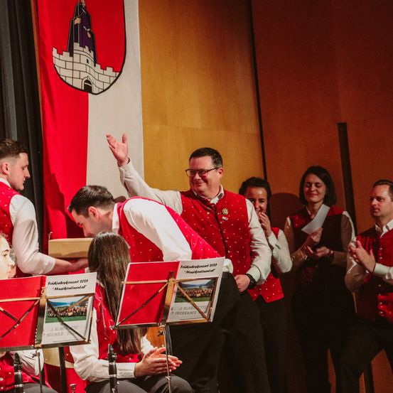 Eine Gruppe von Musikern, sowohl Männer als auch Frauen, tritt auf der Bühne auf. Sie tragen rote Westen und Brillen. Die Frau vorne spielt Flöte, während der Mann neben ihr Saxophon spielt. Hinter ihnen befindet sich eine Fahne und eine Wand. Im Hintergrund klatschen Menschen. Die Notenblätter auf dem Notenständer sind mit 'Freitagabend 2019' betitelt.