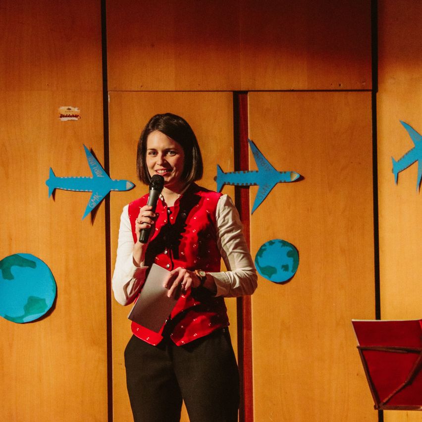 Eine Frau steht auf einer Bühne mit einem Mikrofon in der Hand und hält ein Stück Papier. Sie trägt ein rotes Weste. Hinter ihr befindet sich eine Holzwand mit blauen Flugzeugausschnitten und einem Globus.