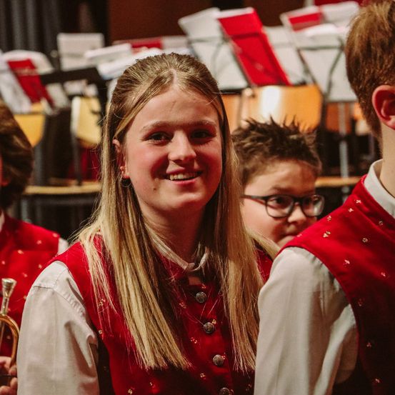 Eine junge Frau mit blonden Haaren lächelt für die Kamera und trägt eine rote Weste mit goldenen Knöpfen. Hinter ihr schauen ein junger Mann mit Brille und eine weitere Person etwas an. Im Hintergrund befinden sich Stühle und Notenständer mit roten und weißen Notenblättern.