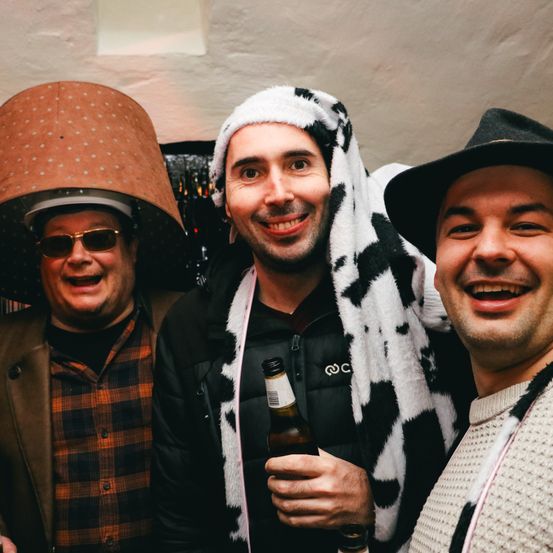 Drei Männer stehen in einem Raum, tragen Hüte, lächeln und schauen zur Kamera. Der Mann in der Mitte hält eine Bierflasche.