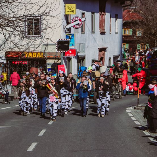 Ein Straßenumzug mit einer Musikkapelle in Kuhkostümen. Einige Menschen sind auf Fahrrädern und andere stehen an der Seite. Im Hintergrund befinden sich Gebäude mit Schildern.