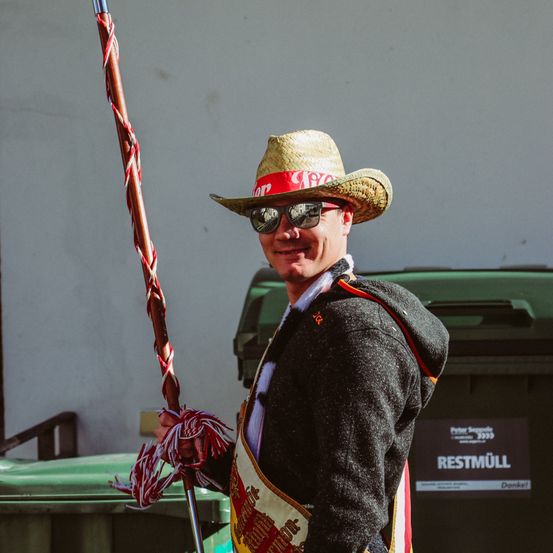 Ein Mann in einem Strohhut mit rotem Band und Sonnenbrille lächelt und hält einen zeremoniellen Stab. Im Hintergrund befindet sich eine Mülltonne mit einem Schild, das Restmuell liest.