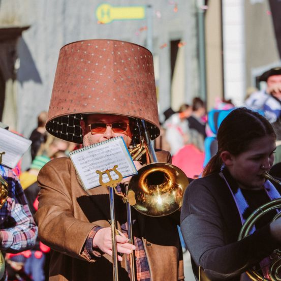 Ein Mann mit einem großen Lampenschirm spielt Posaune, während eine Frau neben ihm eine französische Horn spielt. Sie stehen draußen in einer Menschenmenge.