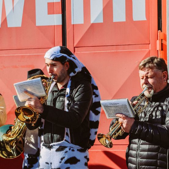 Zwei Männer in Winterkleidung und Kuhkostümen spielen Musikinstrumente. Der Mann auf der linken Seite spielt Trompete, der Mann auf der rechten Seite spielt Posaune.