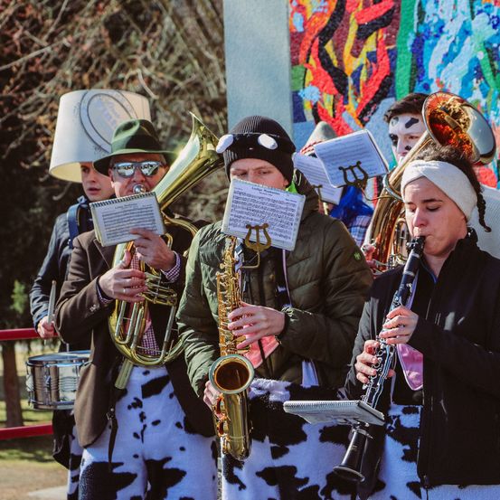 Eine Gruppe von Musikern in Kostümen tritt draußen auf. Sie halten Notenblätter in den Händen und spielen Instrumente. Einer spielt die Posaune, ein anderer das Saxophon und ein weiterer die Klarinette. Hinter ihnen befindet sich eine farbenfrohe Wandmalerei.