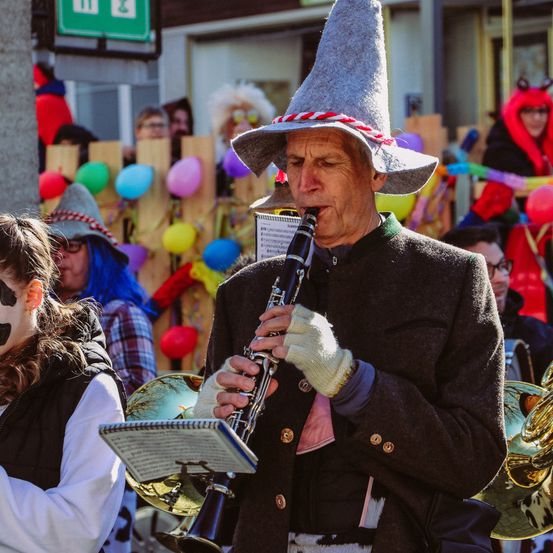 Ein älterer Mann mit einem hohen Hut spielt eine Klarinette bei einer Straßenparade. Dahinter sind andere Teilnehmer in bunten Kostümen und mit Musikinstrumenten zu sehen, darunter eine Tuba.