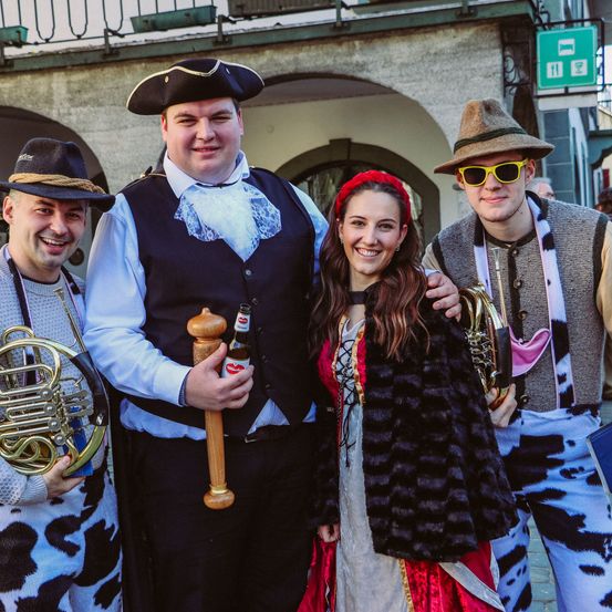 Vier Personen in traditioneller deutscher Kleidung posieren für ein Foto. Der Mann auf der linken Seite hält eine französische Horn, und der Mann in der Mitte hält eine Flasche. Die Frau in der Mitte trägt eine rote Kopfbinde und einen Pelzmantel.