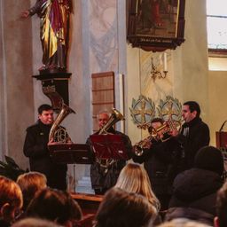Ein Blechbläserensemble aus vier Männern spielt in einer Kirche mit einer goldenen Statue im Hintergrund. Sie spielen Trompeten und Tuben. Ein Publikum sitzt vor ihnen.