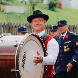 Ein Mann spielt eine große Trommel und lächelt, während ein weiterer Mann in Uniform hinter ihm steht. Sie sind draußen im Regen, in der Nähe eines Zauns.