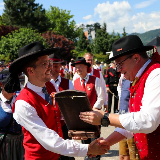 Ein Mann in einem roten Weste mit einem roten Hut schüttelt die Hand eines anderen Mannes, der einen schwarzen Hut und eine Uhr trägt. Sie halten eine Holzkiste. Im Hintergrund stehen einige Leute und schauen sie an.
