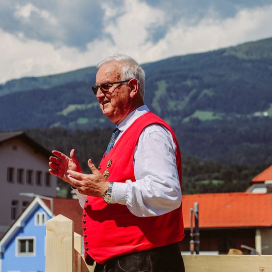 Ein älterer Mann in einem roten Vest steht vor einem Holzzaun und spricht. Im Hintergrund befinden sich Häuser und Berge unter einem teilweise bewölkten Himmel.
