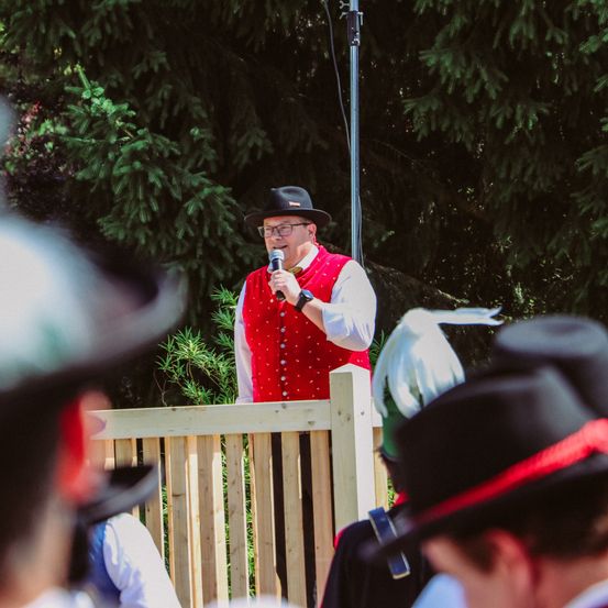 Ein Mann in einem roten Weste und Hut spricht in ein Mikrofon vor einem Holzzaun. Menschen mit schwarzen Hüten hören hinter dem Zaun zu.