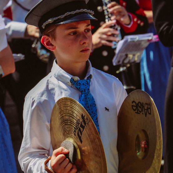 Ein junger Junge in einem weißen Hemd und einer blauen Krawatte hält zwei Becken und schaut in die Kamera. Hinter ihm spielt eine Person Flöte und eine andere Person hält einen Notenständer.