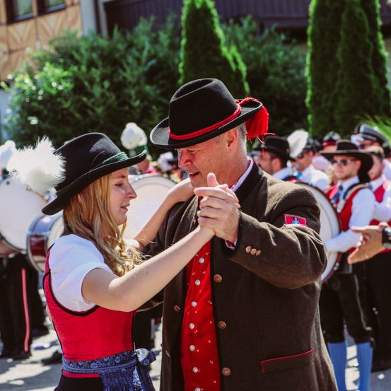 Ein Mann und eine Frau tanzen vor einer Marschkapelle, beide in traditioneller Kleidung. Die Frau trägt ein rotes und weißes Kleid, der Mann einen braunen Mantel. Dahinter spielen Mitglieder der Band Trommeln und andere Instrumente und tragen Uniformen.