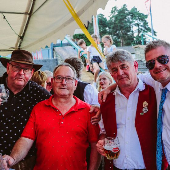 Drei Männer stehen eng beieinander, zwei von ihnen halten Gläser mit Bier in der Hand und lächeln in die Kamera. Im Hintergrund spielen zwei Kinder auf einer Rutsche. Bäume und ein weißes Zelt sind sichtbar.
