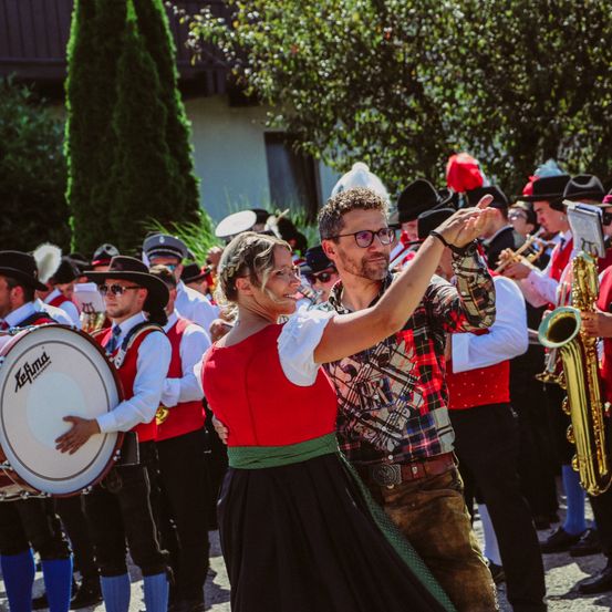 Ein Paar in traditioneller bayerischer Kleidung tanzt vor einer Marschkapelle, umgeben von anderen in ähnlicher Kleidung, an einem sonnigen Tag.