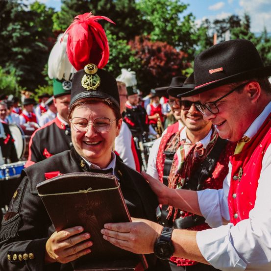Ein Mann hält ein großes Buch, trägt eine Uniform mit Hut und lächelt. Ein Mann in einem roten Vest und Hut steht neben ihm. Dahinter stehen mehrere Menschen in Hüten und Uniformen, die möglicherweise an einem Umzug teilnehmen. Im Hintergrund sind Bäume und eine Trommel zu sehen.