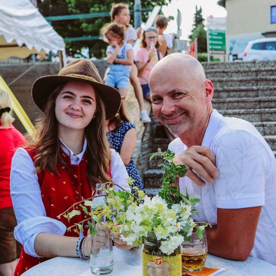 Ein Mann und eine Frau sitzen lächelnd an einem Tisch. Die Frau trägt einen Hut und der Mann ein weißes Hemd. Sie halten ein Glas mit einer Blume darin. Hinter ihnen stehen mehrere Personen auf den Stufen.