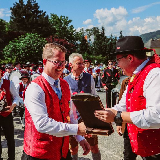Ein Mann mit einem Zertifikat schaut einen anderen Mann an, der eine kleine Schachtel hält und lächelt, während andere in roten Westen und Hüten hinter ihnen stehen.