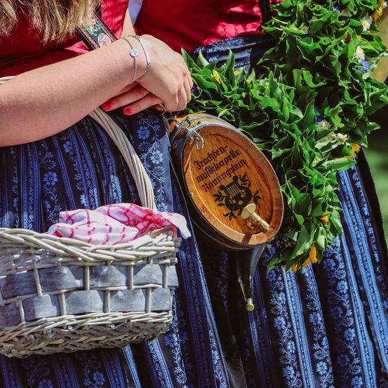 Eine Frau in einem blau-weißen traditionellen Kleid mit einem weißen Weidenkorb und einer runden Ledertasche mit dem Wort 'Trachten' darauf.