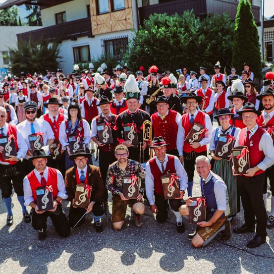 Eine große Gruppe von Menschen in roten Westen und Mützen posiert für ein Foto. Einige halten Auszeichnungen. Dahinter steht ein Gebäude mit Bäumen und einem Berg in der Ferne.