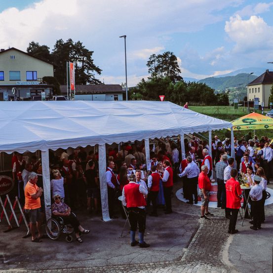 Viele Menschen versammeln sich unter einem weißen Zelt und tragen rote und weiße Hemden. Einige stehen, andere sitzen auf Stühlen. Im Hintergrund sind auch Menschen zu sehen, in der Nähe eines Gebäudes mit der Aufschrift "WaffenDoc".