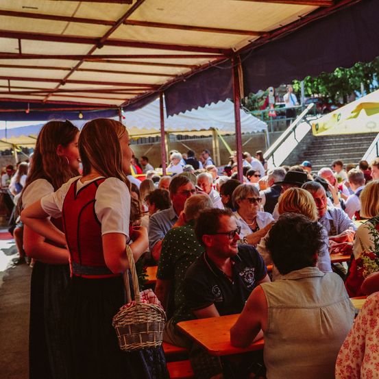 Eine Gruppe von Menschen sitzt an Tischen in einem Restaurant im Freien. Eine Frau in traditioneller deutscher Kleidung steht mit einem Korb in der Hand. Einige der Menschen tragen Brillen und Hüte. Im Hintergrund ist eine Treppe zu sehen.