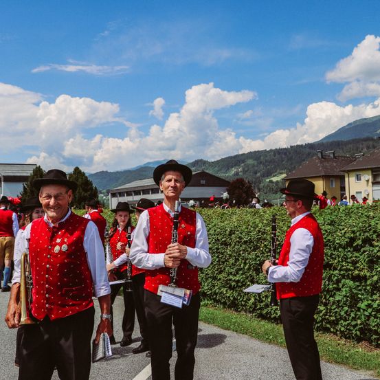 Eine Gruppe von Musikern in roten Westen, weißen Langarmhemden und schwarzen Hosen steht vor einer Hecke. Sie spielen ihre Instrumente, darunter Trompete, Klarinette und Posaune. Dahinter befinden sich einige Gebäude, darunter ein Haus mit braunem Dach und einem Turm.