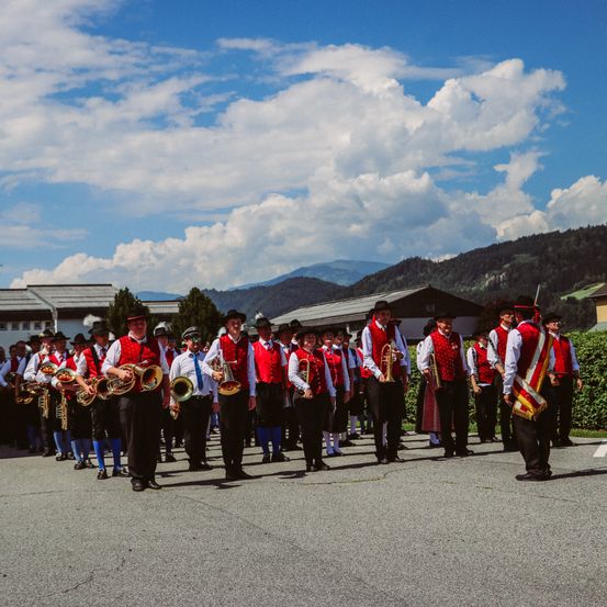 Eine Gruppe von Männern in roten Westen und schwarzen Hosen spielt Blechblasinstrumente im Freien. Sie stehen in einer Reihe mit einigen Leuten vor ihnen. Dahinter befinden sich Häuser und Berge.