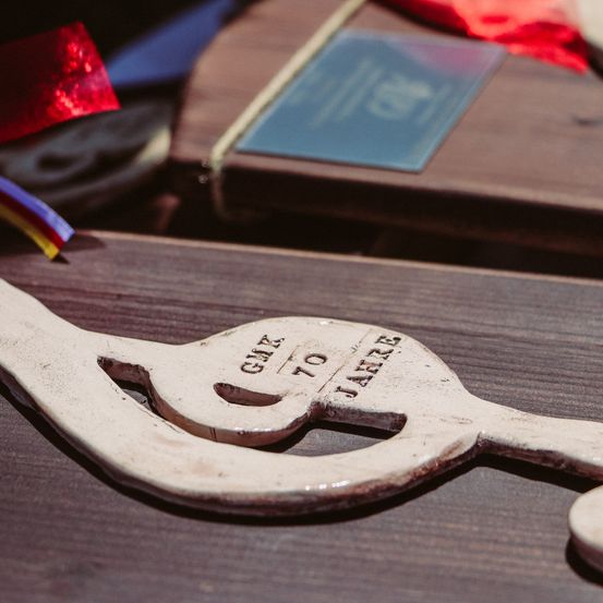 Ein Nahaufnahme eines Vintage-Gitarren-Picks mit der Gravur 'Gail 70 zu Jare' auf einer Holzoberfläche. Der Pick wird von anderen Holzgegenständen begleitet, einschließlich einer Medaille und Bändern, die im Hintergrund leicht verschwommen sind.