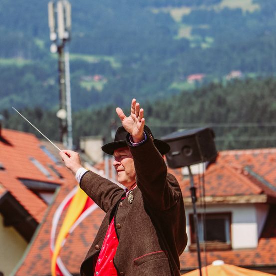 Ein Mann in einem roten Westchen und einem schwarzen Hut hebt seine Hände und wedelt mit einem Taktstock, während er im Hintergrund eine Fahne hält.