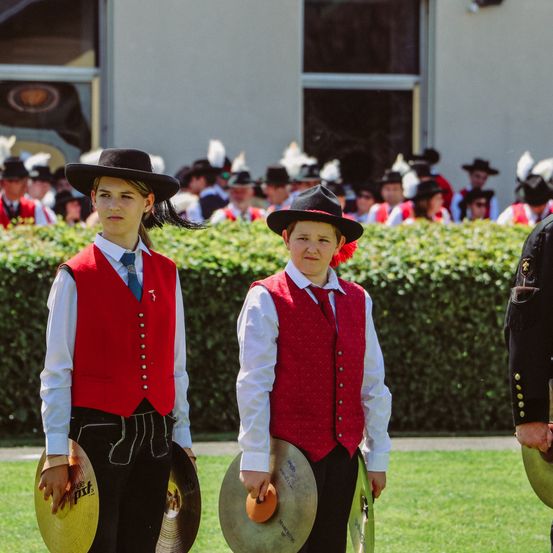 Zwei junge Jungen in roten Westen und Hüten mit rotem Rand halten Becken und stehen vor einer Menschenmenge in ähnlichen Uniformen.