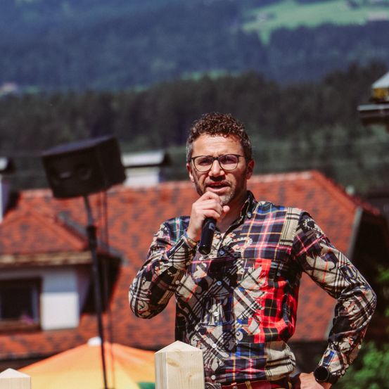 Ein Mann in einem karierten Hemd spricht in ein Mikrofon vor einem Gebäude mit rotem Dach.