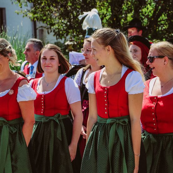 Eine Gruppe von Frauen in traditioneller deutscher Kleidung steht in einer Reihe, nach vorne gerichtet, während die Menschen hinter ihnen unscharf sind.