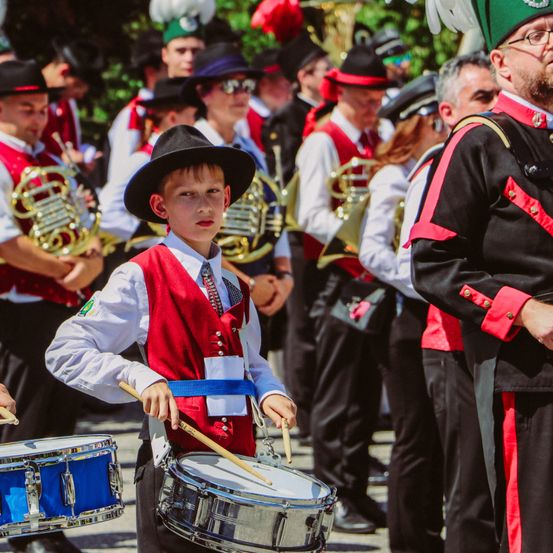 Ein Junge, der Trommeln spielt, ist von einer Band umgeben, mit Musikern, die französische Hörner und andere Instrumente spielen, in traditionellen Uniformen gekleidet. Der Hintergrund ist unscharf und konzentriert sich auf den Jungen und seine Trommel.