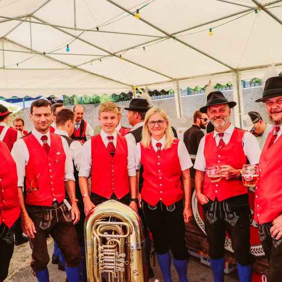 Eine Gruppe von Menschen in traditioneller deutscher Kleidung steht unter einem Zelt, hält Biergläser in den Händen und lächelt. Ein großer Tuba steht im Vordergrund.